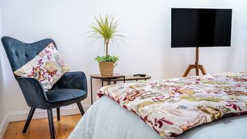 Une chambre à coucher avec un lit, une chaise, une petite table avec une plante et un téléviseur posé sur un meuble.