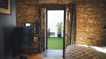 A room with a stone wall, a bed, a desk with a chair, and an open door leading to a small outdoor area with a plant.