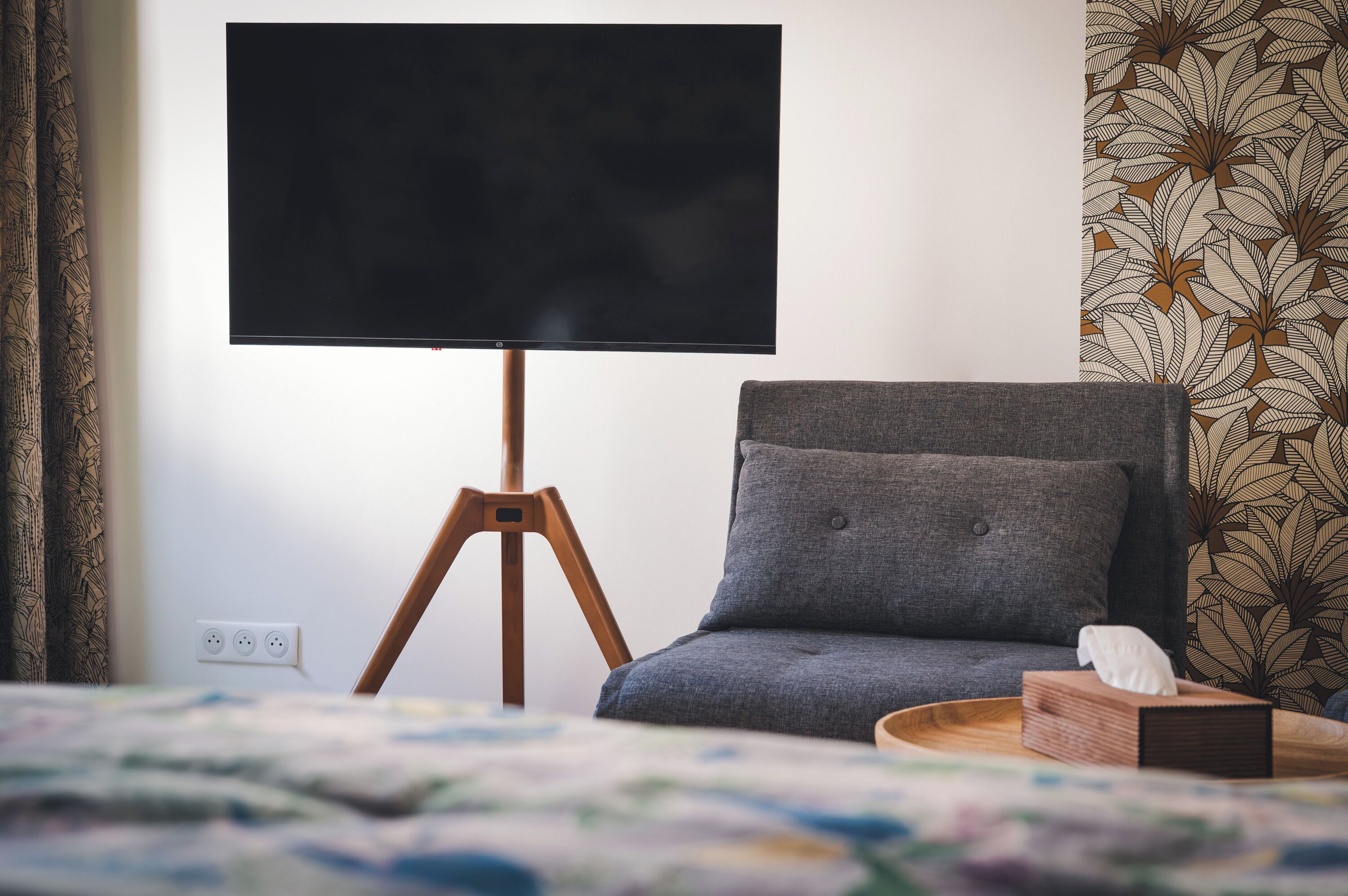 Een kamer met een bed, een grijze fauteuil, een houten nachtkastje, een televisie op een statief en behang met een patroon.
