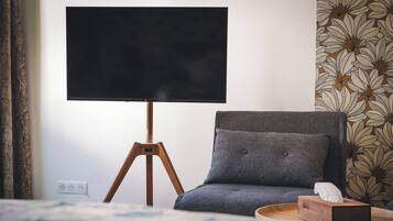 Een kamer met een bed, een grijze fauteuil, een houten nachtkastje, een televisie op een statief en behang met een patroon.