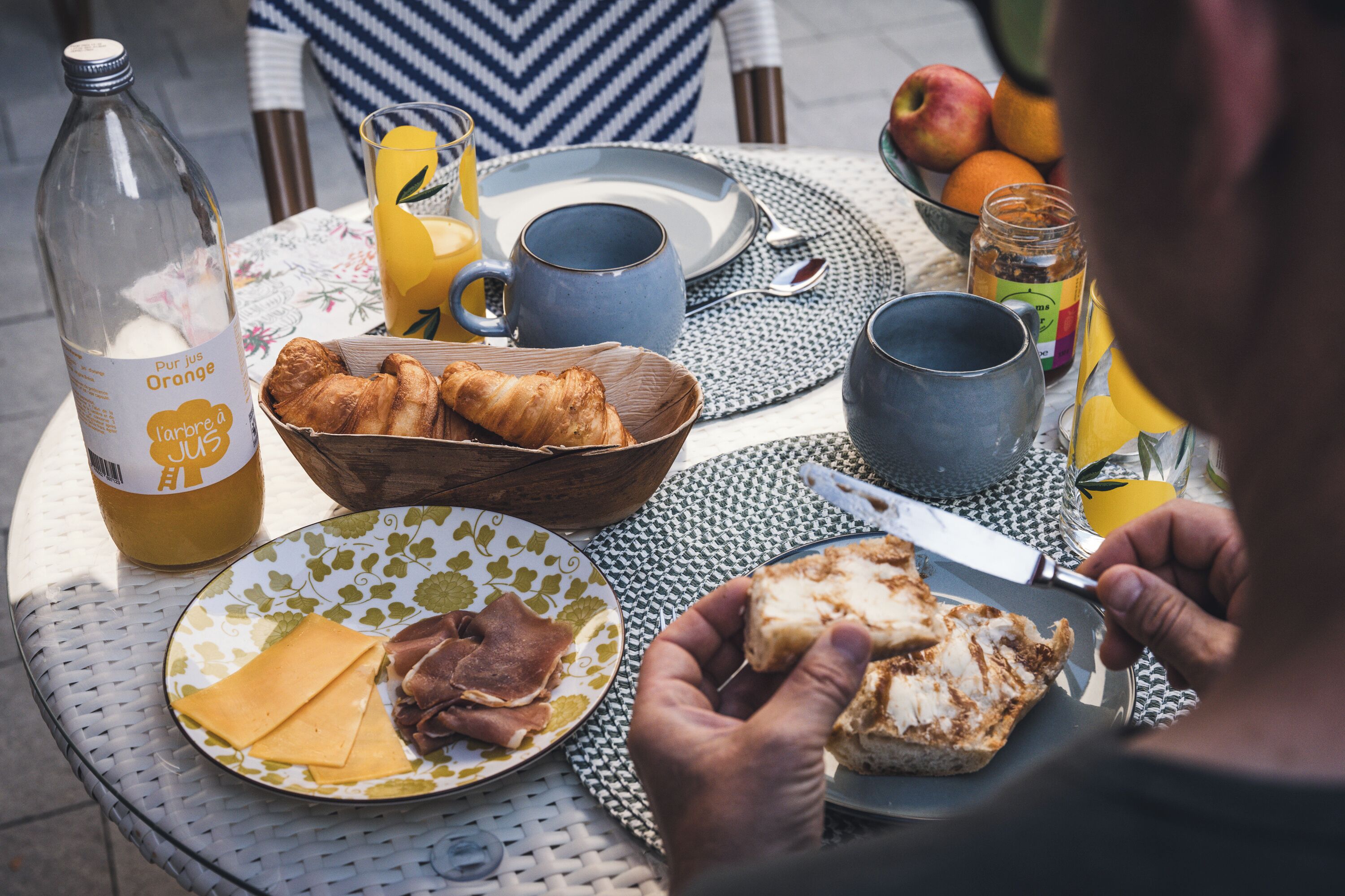 Desayuno con croissants, queso, jamón y tostadas sobre una mesa con una botella, tazas y un bol de fruta.