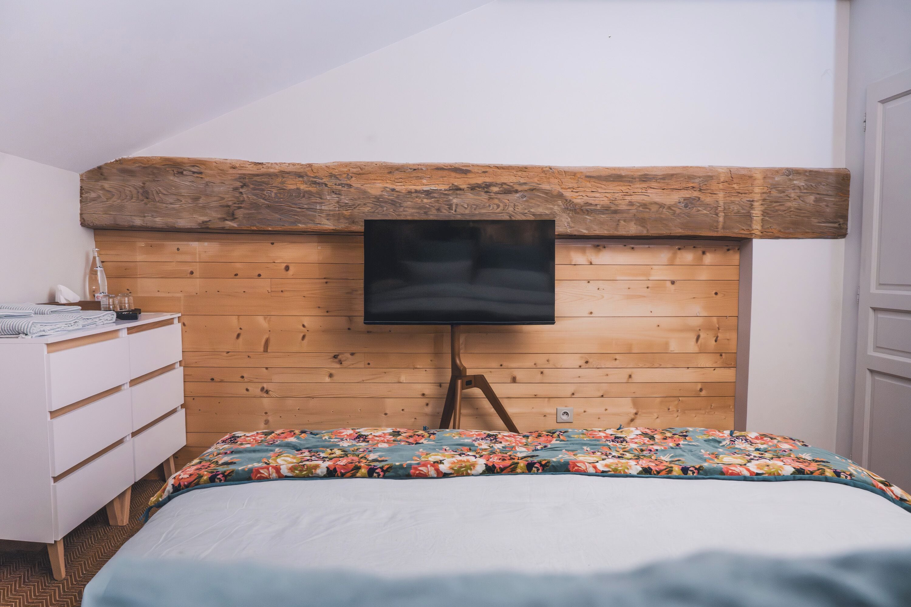 A bedroom with a wooden headboard, a bed with a floral bedspread, a white dresser, and a flat-screen TV mounted on the headboard.