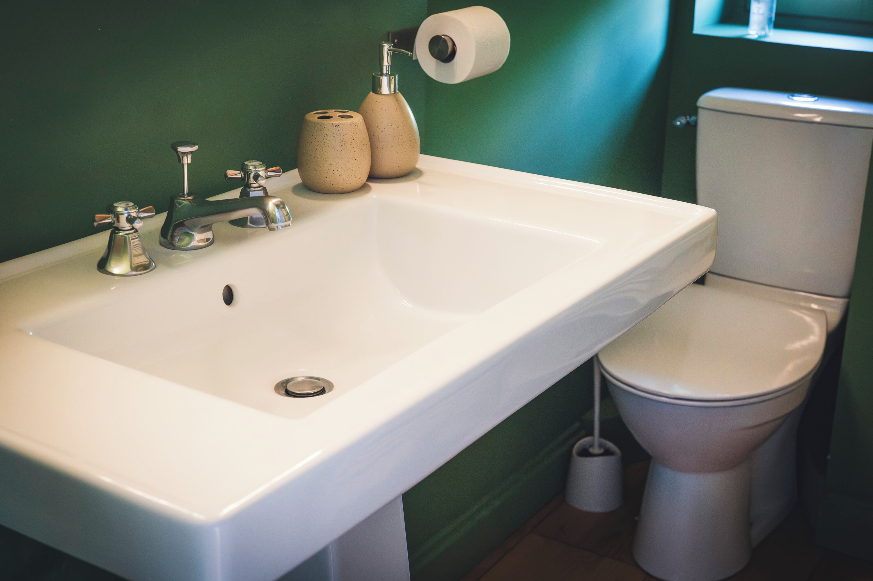 Ein Badezimmer mit einem weißen Waschbecken, zwei Wasserhähnen, einer Toilette und einer Toilettenpapierrolle an der Wand.