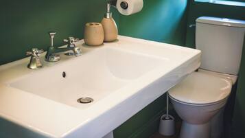 Un baño con un lavamanos blanco, dos grifos, un inodoro y un rollo de papel higiénico en la pared.