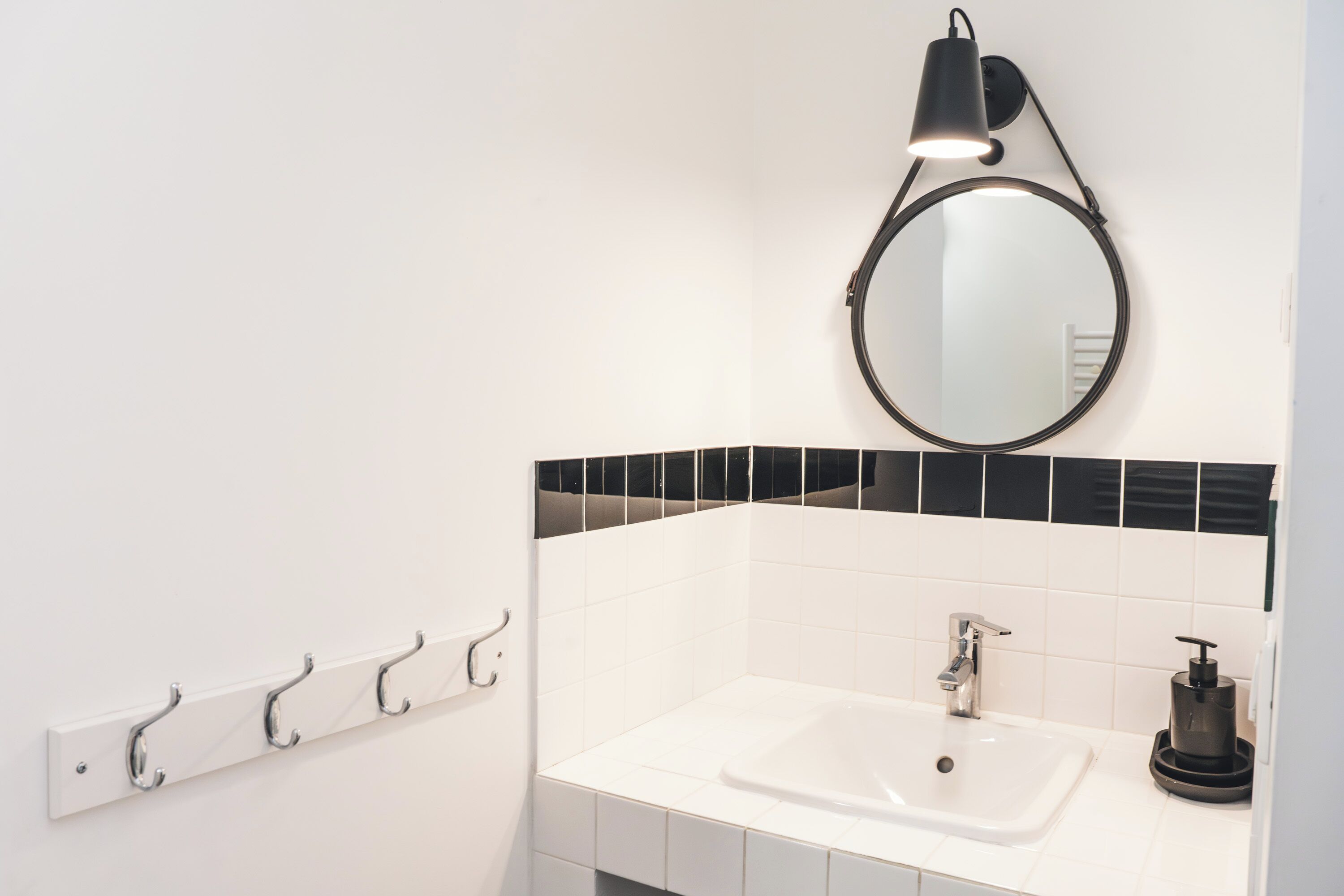 A bathroom with a white sink, black-tiled backsplash, a round mirror with a black light, and a wall-mounted towel rack.
