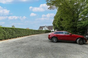 Une voiture rouge est garée sur une allée de gravier, avec une haie d’un côté et une maison en arrière-plan.