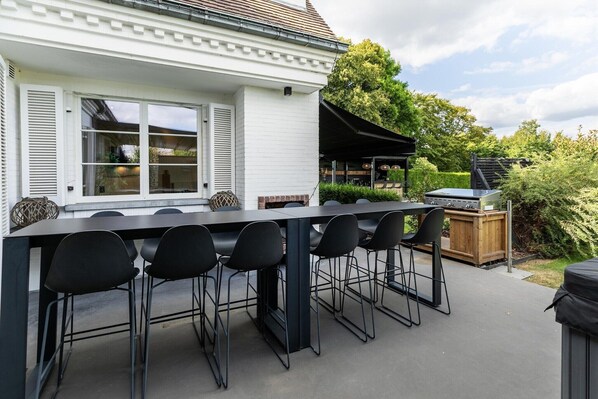 Un espace extérieur aménagé pour les repas, avec une longue table et plusieurs chaises, un barbecue et un coin salon abrité.