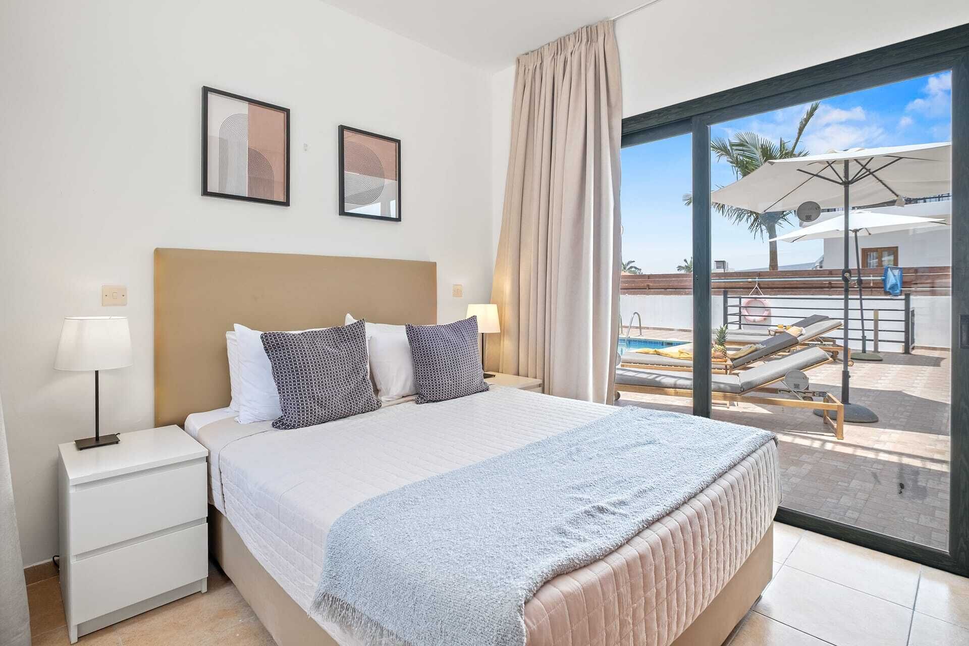 A hotel room with a bed, a nightstand, a lamp, and a view of a pool area.