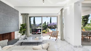 A modern living room with a sofa, a coffee table, and a TV. There is a sliding glass door leading to a balcony with a pool and garden. - Akamé Luxury Beach Villa by Nomads (Paphos)