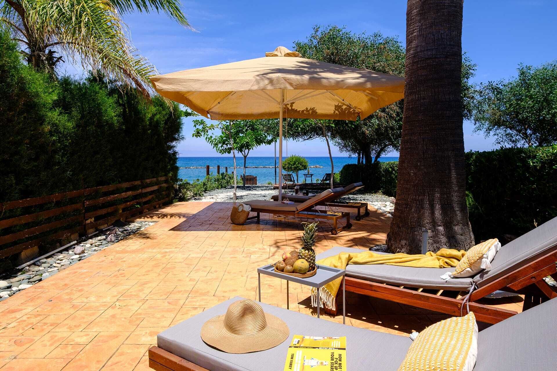 A beachside lounge area with sun loungers, a table with fruits, and a large umbrella.