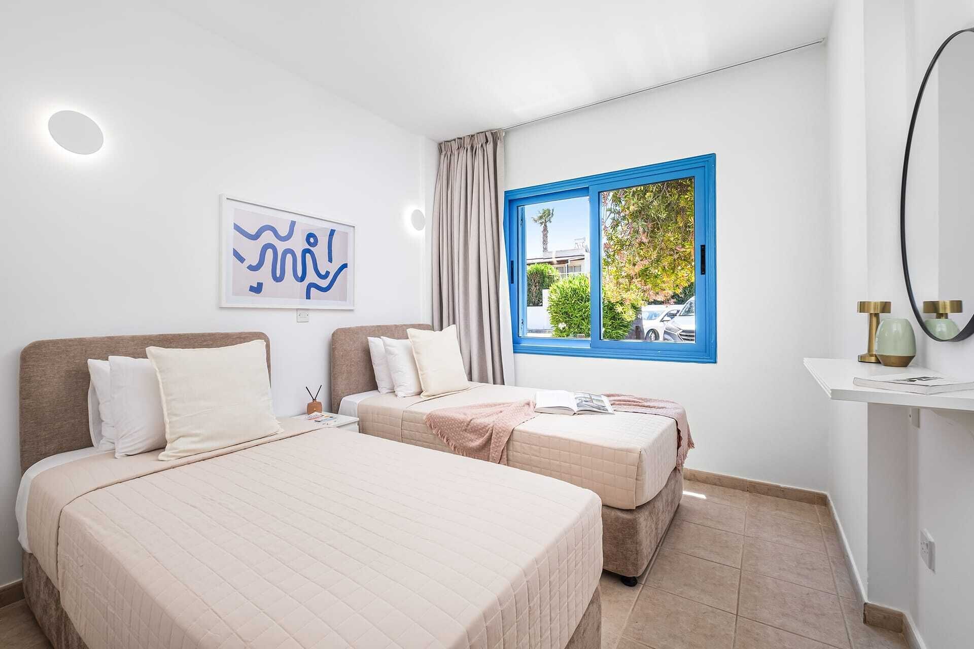 Une chambre avec deux lits, une fenêtre à cadre bleu, une œuvre d’art accrochée au mur et une table de chevet avec une lampe.