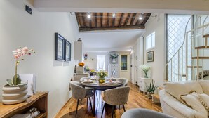Uma área de refeições com uma mesa redonda, cadeiras e um vaso de flores. Há um sofá e uma estante em primeiro plano.