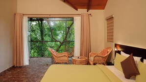 A hotel room with a bed, two wicker chairs, a small table, and a view of lush greenery through a glass door. - Panchavadi Farm Stay (Kumily)