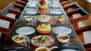 A long dining table set for a meal with various dishes, including rice, salad, and a fish dish, surrounded by chairs. - Panchavadi Farm Stay (Kumily)