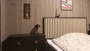 Un dormitorio con pared de madera, una cama con un cojín estampado, una mesita de noche con una lámpara y un cuadro con una imagen de un esquiador.