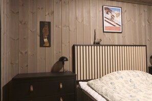 Un dormitorio con pared de madera, una cama con un cojín estampado, una mesita de noche con una lámpara y un cuadro con una imagen de un esquiador.