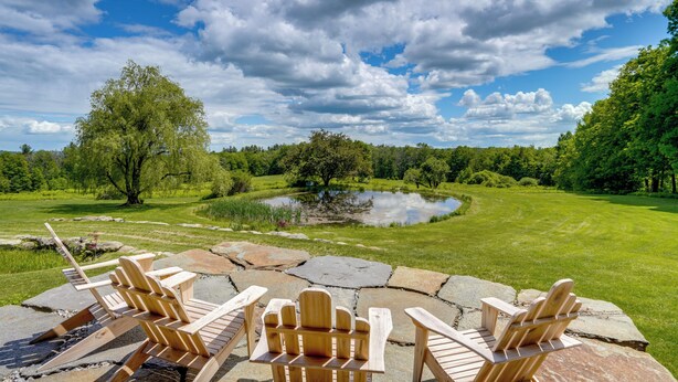 Eine Steinterrasse mit Adirondack-Stühlen mit Blick auf einen Teich und eine grüne Landschaft.