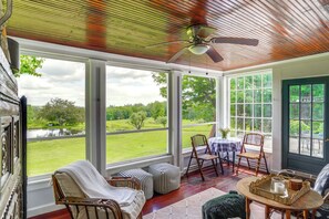 Ein Wintergarten mit Deckenventilator, großen Fenstern, Korbmöbeln und Blick auf eine grüne Landschaft.