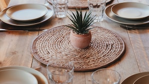 Uma mesa de jantar de madeira posta com pratos, copos e plantas em vasos.