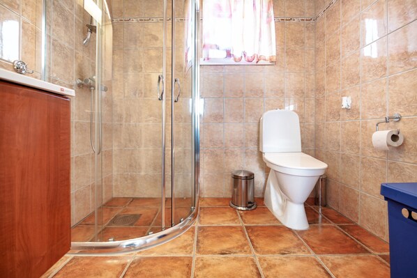 Un baño con cabina de ducha de vidrio, un inodoro, un cubo de basura y otro cubo de basura azul.