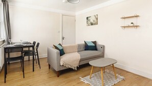 Una sala de estar con un sofá gris, una mesa de comedor con sillas, una pequeña mesa redonda, una lámpara colgante verde y una puerta blanca.