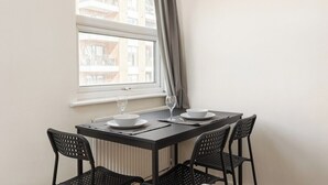 Un comedor con una mesa preparada para dos personas, dos sillas, un ventanal con vistas a edificios y un radiador debajo de la ventana.
