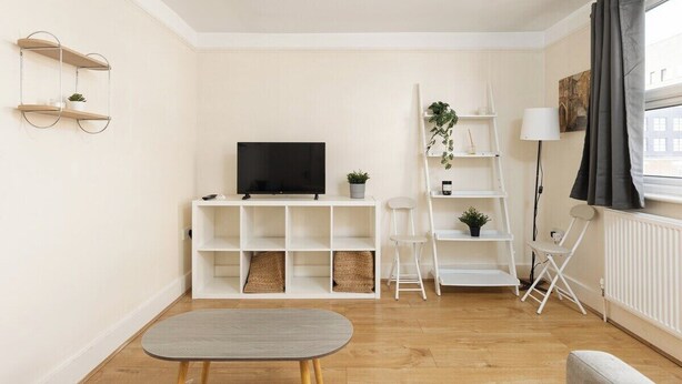 Una sala de estar moderna con televisor de pantalla plana, estanterías blancas, una mesita y un ventanal con cortinas.