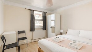 Un dormitorio con cama, una silla, un radiador, una ventana con cortinas y una mesita de noche.