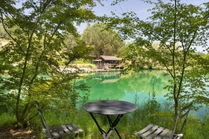 Blick auf ein Seeufer mit einer Holzterrasse, einem kleinen Gebäude und einem Tisch mit Stühlen.