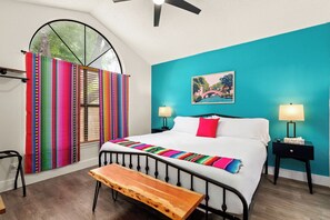 Ein Schlafzimmer mit einem Bett, zwei Nachttischlampen, einer Bank, einem Fenster mit Vorhängen und einem Gemälde an der Wand.