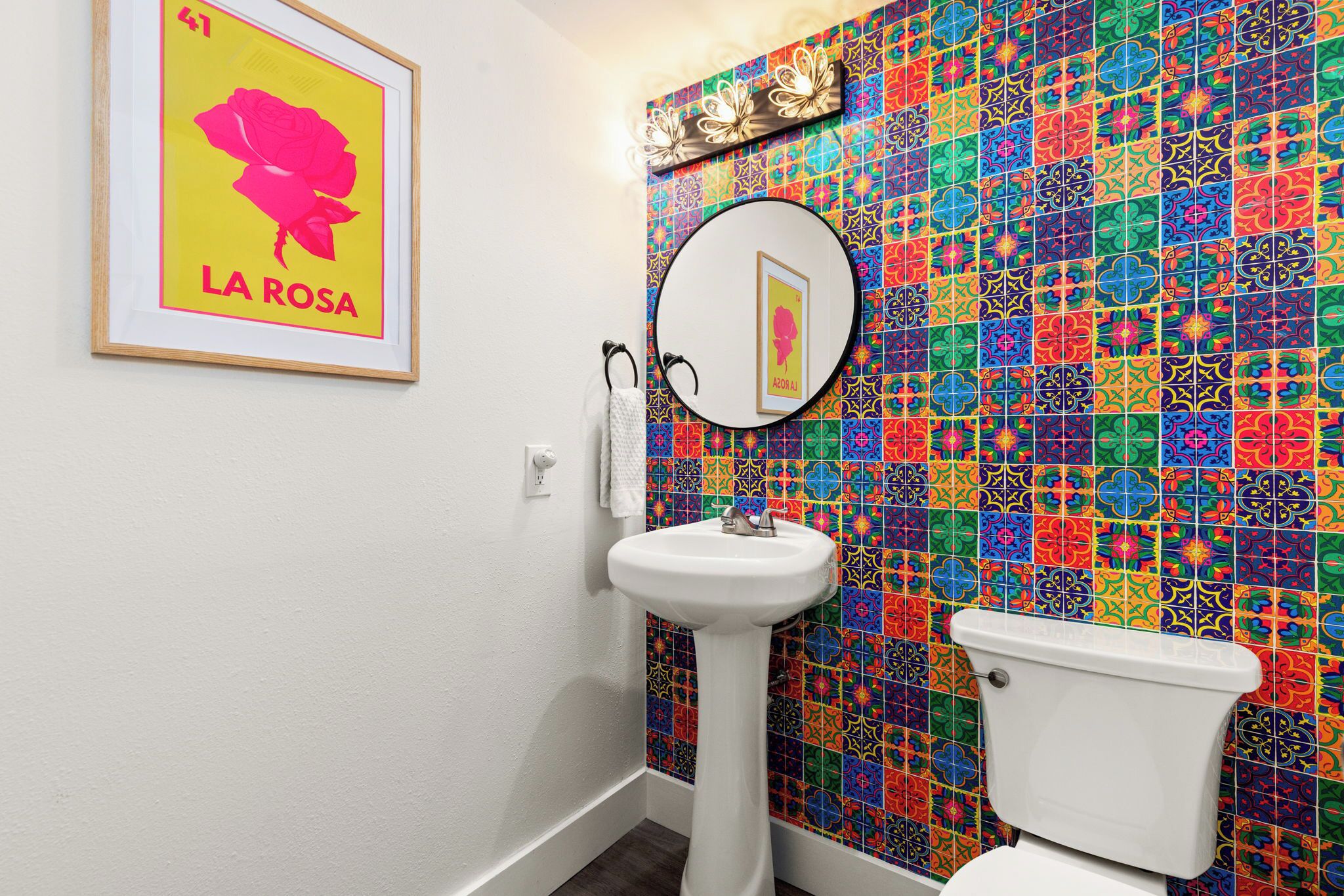 Une salle de bain avec un mur recouvert de carreaux colorés, une affiche encadrée, un lavabo et des toilettes.
