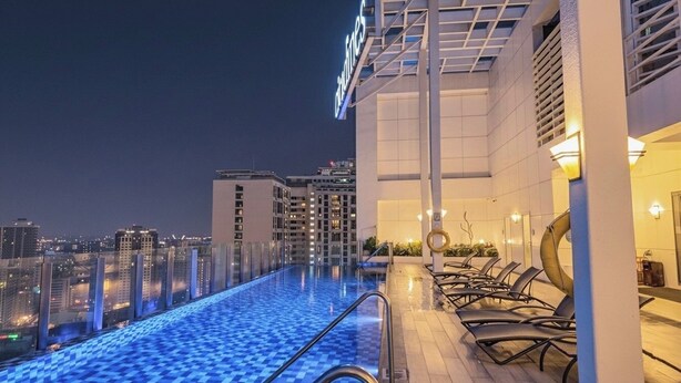 En infinitypool med utsikt över staden, solstolar och en modern byggnadsfasad.
