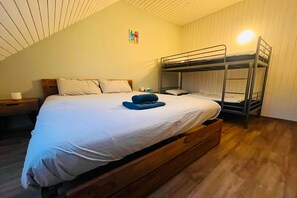 Une chambre avec un lit superposé, un plancher en bois, un lit simple avec une literie blanche, une table de chevet et une lampe fixée au mur.