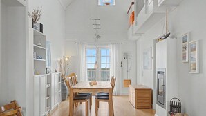En rummelig spiseplads med et træbord og stole, et hvidt skab med hylder, et vindue med gardiner og udsigt udad.