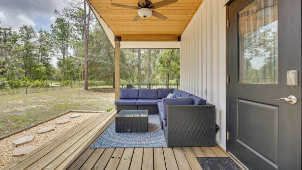 A covered porch with a ceiling fan, a dark-colored door, and a blue outdoor seating area. - 6 Mi to Three Sisters Springs! Extended Getaway (Homosassa)