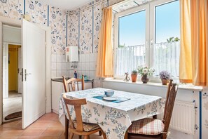 A kitchen with a dining table, chairs, a window with curtains, and a view of greenery outside. - Holiday Apartment for 3 Guests (45 m²) in Brunsbüttel (Brunsbüttel)