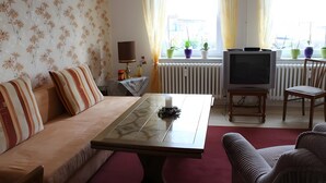 Una sala de estar con un sofá, una mesa de centro, un televisor sobre un mueble bajo y plantas en macetas junto a la ventana.