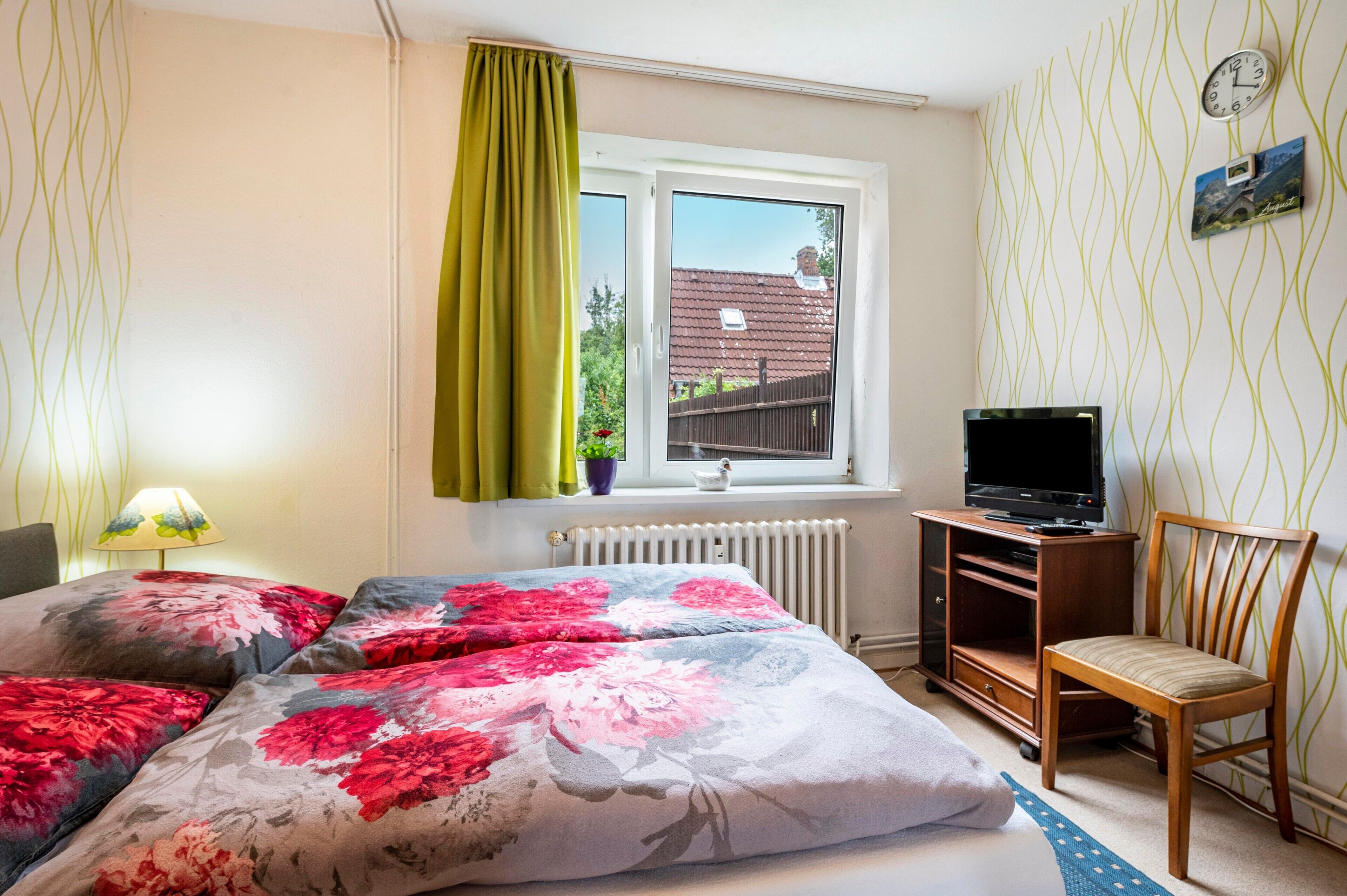 A bedroom with a bed, a TV, a chair, a window, and a radiator.