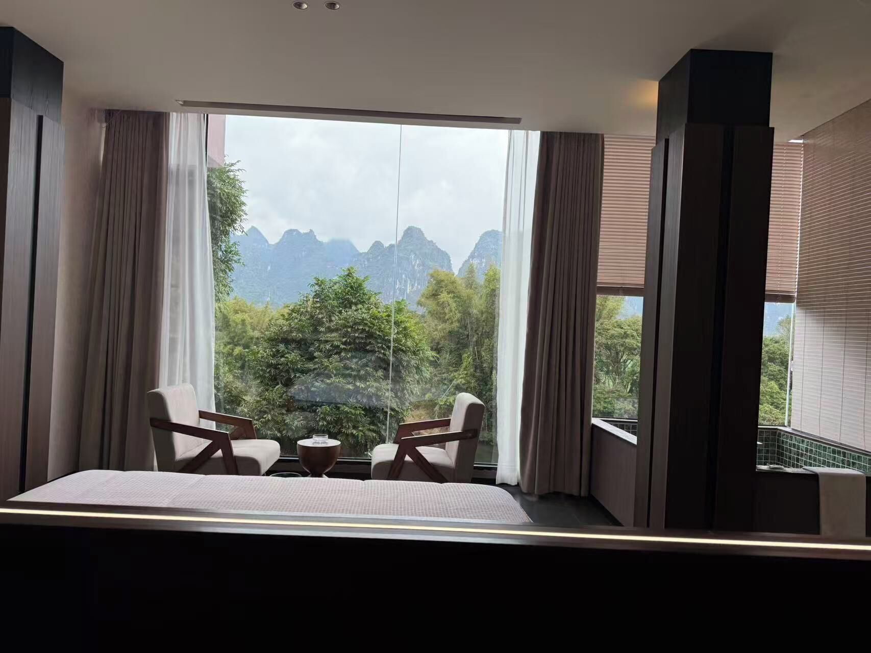 A hotel room with a large window, two chairs, a small table, and a bed.