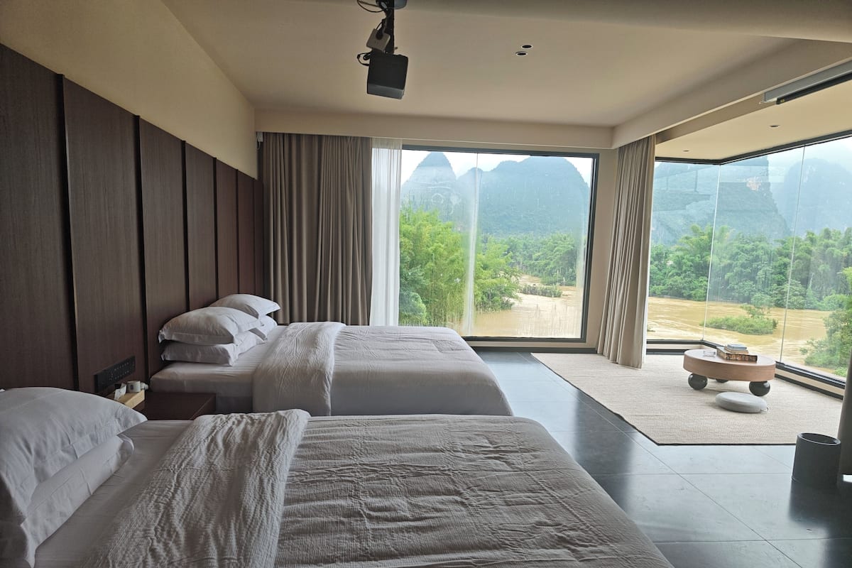Habitación de hotel con dos camas, un ventanal grande, un proyector y una mesita.