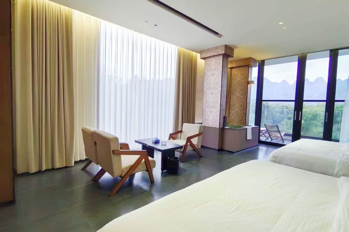 Habitación de hotel con dos camas, un escritorio, sillas y vistas a la montaña, además de amplios ventanales.
