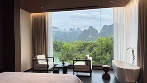 A modern hotel room with a large bed, two armchairs, a small table, a bathtub, and a view of a mountainous landscape.