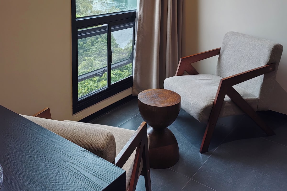 Una habitación con un ventanal grande, una silla, una mesa redonda de madera y vistas a montañas y vegetación.