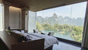 A modern hotel room with a large bed, a desk, and a view of a lake and mountains.