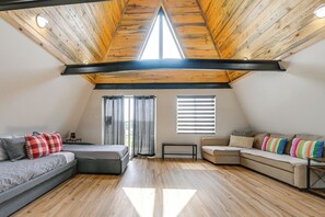 A loft bedroom with a sloped ceiling, wooden beams, a gray sectional sofa, a beige sofa, a small table, and a bench.