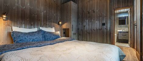A bedroom with a wooden wall, a bed with patterned bedding, and a mirror in the adjacent room.