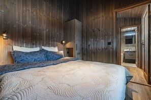 A bedroom with a wooden wall, a bed with patterned bedding, and a mirror in the adjacent room. - Hemsedal ski in/ski out cabin – modern comfort by the slopes (Hemsedal)