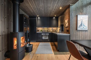 A modern kitchen with a wood-burning stove, black cabinets, and a dining area with a table and chair. - Hemsedal ski in/ski out cabin – modern comfort by the slopes (Hemsedal)
