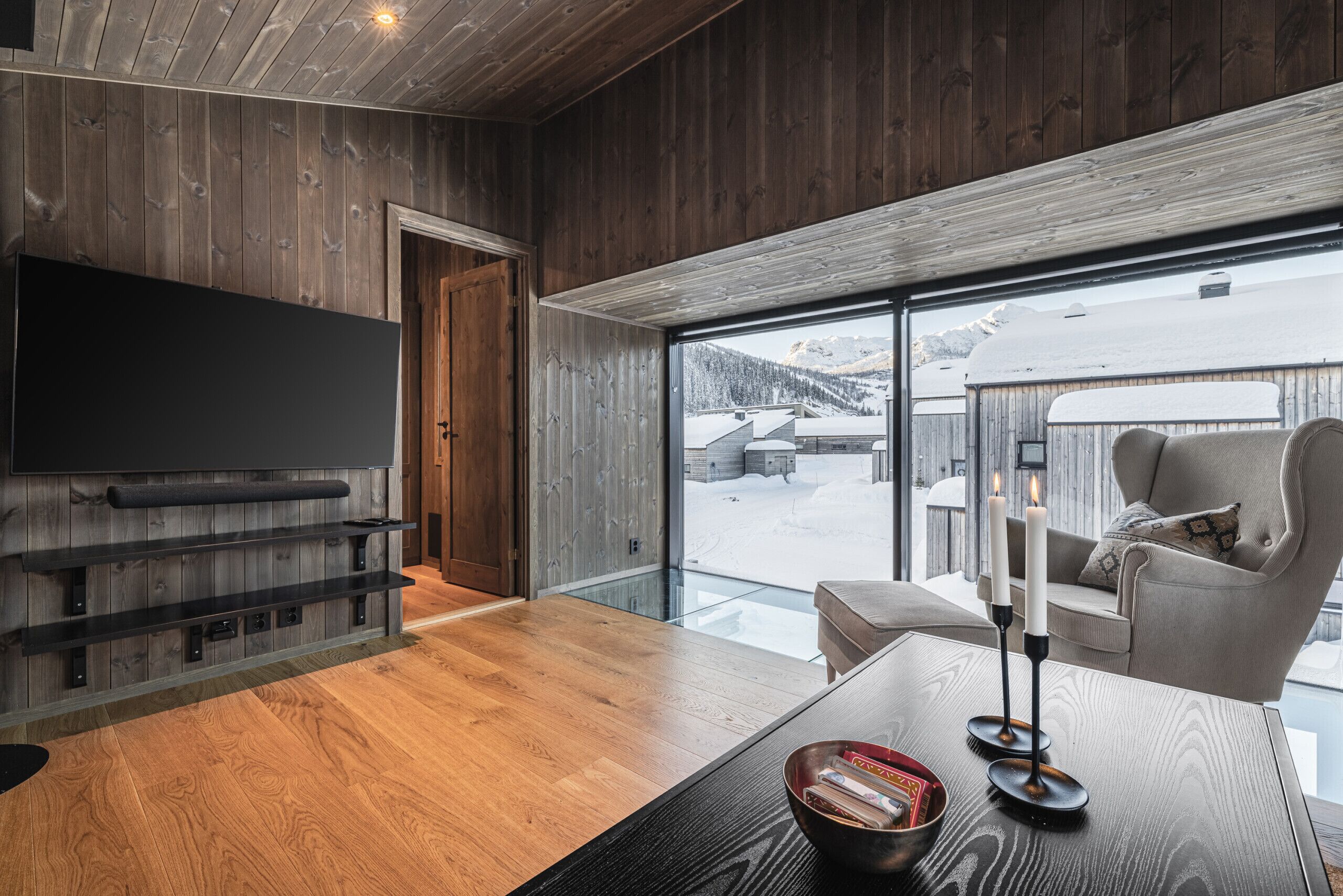 Un intérieur moderne avec un mur en bois, un téléviseur à écran plat, une porte vitrée donnant sur l’extérieur et un coin salon avec une table et des bougies.