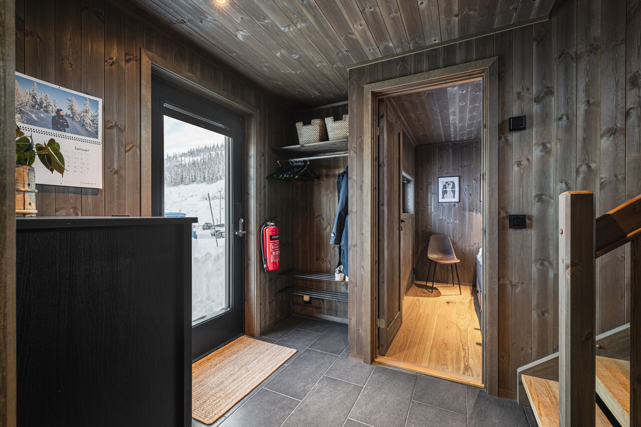 A wooden interior with a door leading to a snowy view, a fire extinguisher, and a chair.